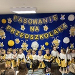 Dzieci w przebraniu za pszczółki stoją na tle dekoracji z okazji pasowania na przedszkolaka